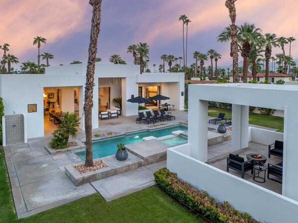 A modern outdoor pool area with lounge chairs, a fire pit, and a view of a residential neighborhood.