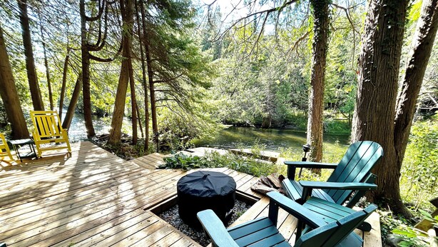 A wooden deck with green Adirondack chairs and a covered seating area, surrounded by trees and a small body of water. - Custom River Front Property on 10 Acres, Sleeps 19 (Traverse City)