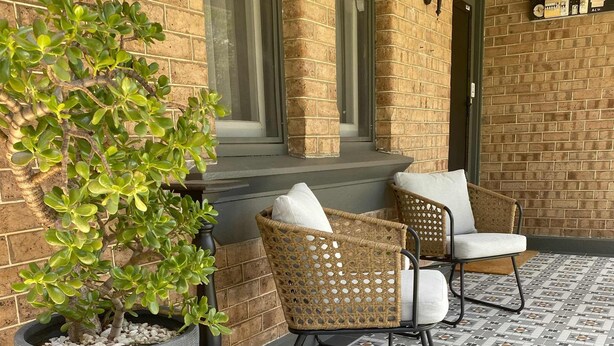 Un bâtiment en briques avec un patio carrelé, deux fauteuils en osier avec des coussins et une grande plante en pot.