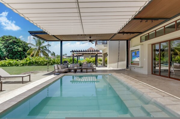 A modern outdoor pool area with a covered seating area, a pergola, and a view of a tropical landscape.
