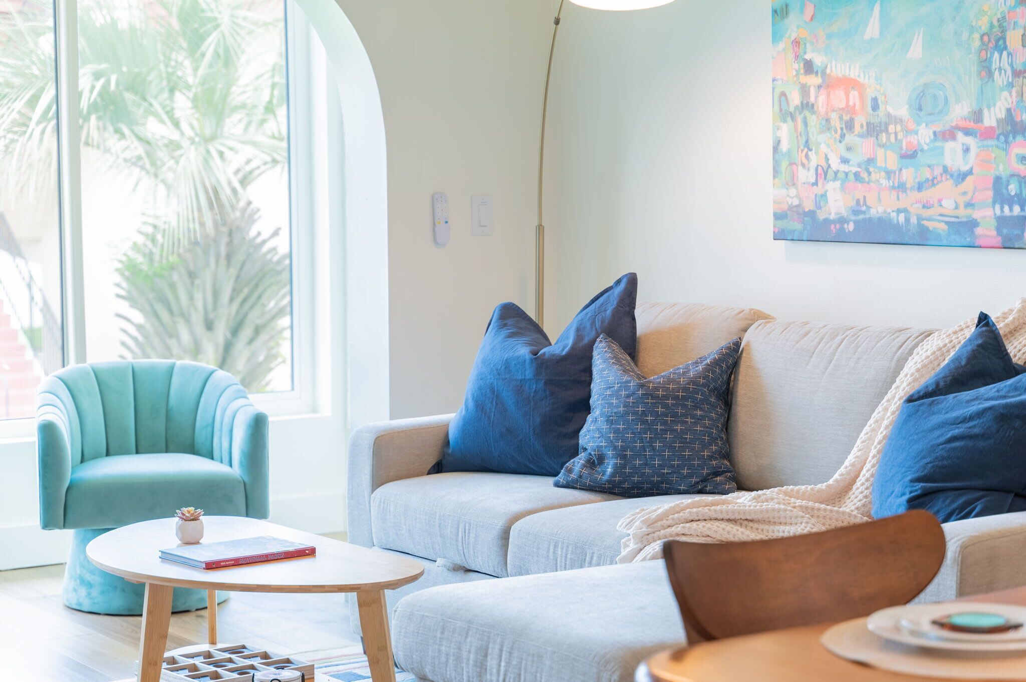Un salon moderne avec un canapé gris clair, des coussins bleus à motifs, une table basse ronde en bois et un fauteuil turquoise.