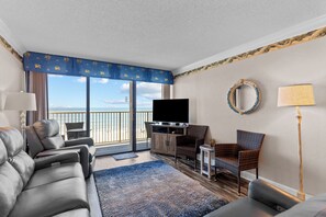 A hotel room with a balcony, a sofa, a chair, a TV, and a lamp. - Oceanfront Condo with Indoor Pool/Hot tub Access (Carolina Beach)