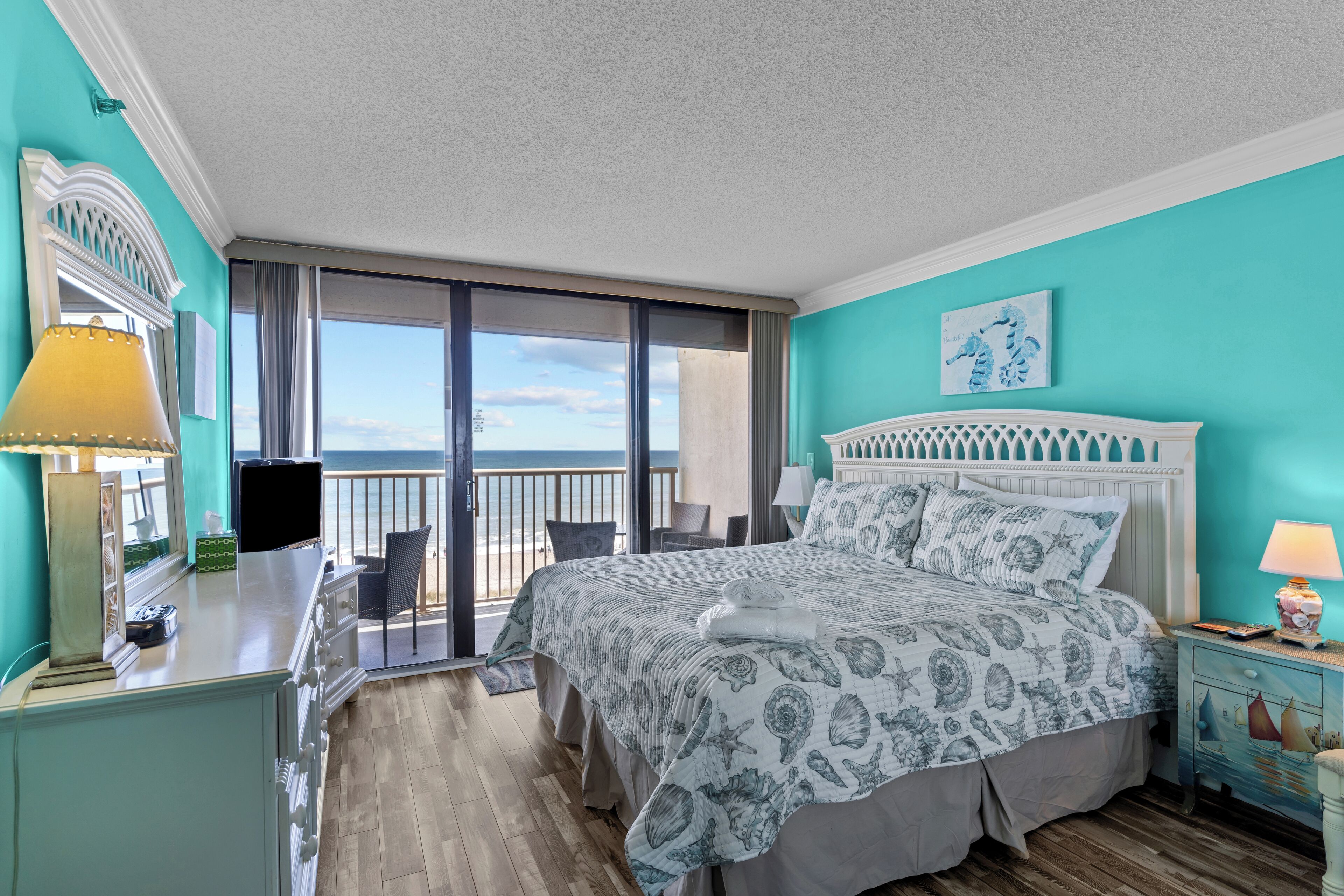 A bedroom with a bed, a desk, a chair, a nightstand, and a view of the beach.
