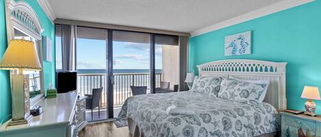 A bedroom with a bed, a desk, a chair, a nightstand, and a view of the beach.