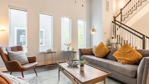 Un salon moderne avec un canapé, un fauteuil et une table basse. On y trouve de grandes fenêtres, un escalier et une petite table d’appoint sur laquelle est posée une plante.