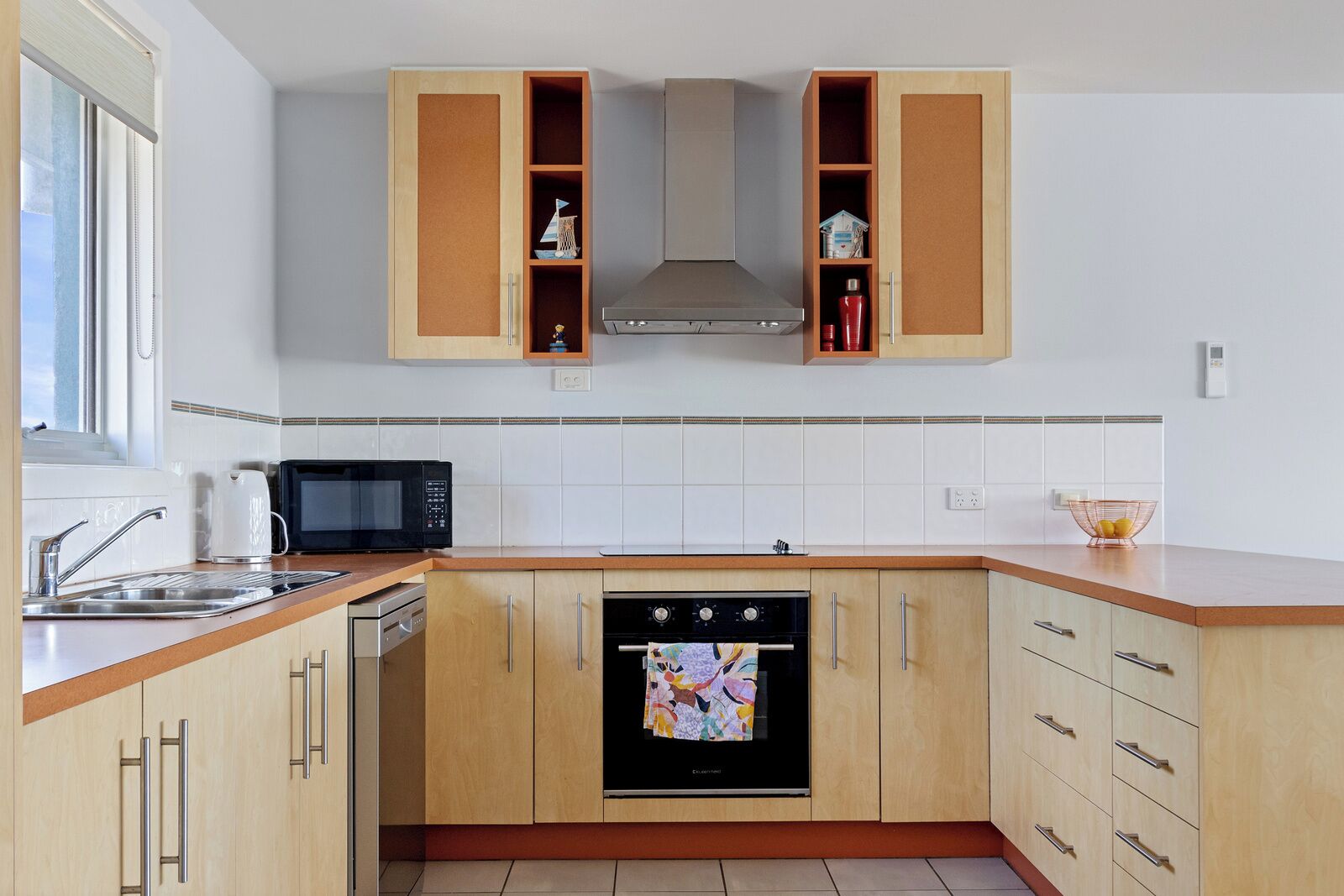 A modern kitchen with wooden cabinets, a built-in oven, a microwave, and a window with blinds.