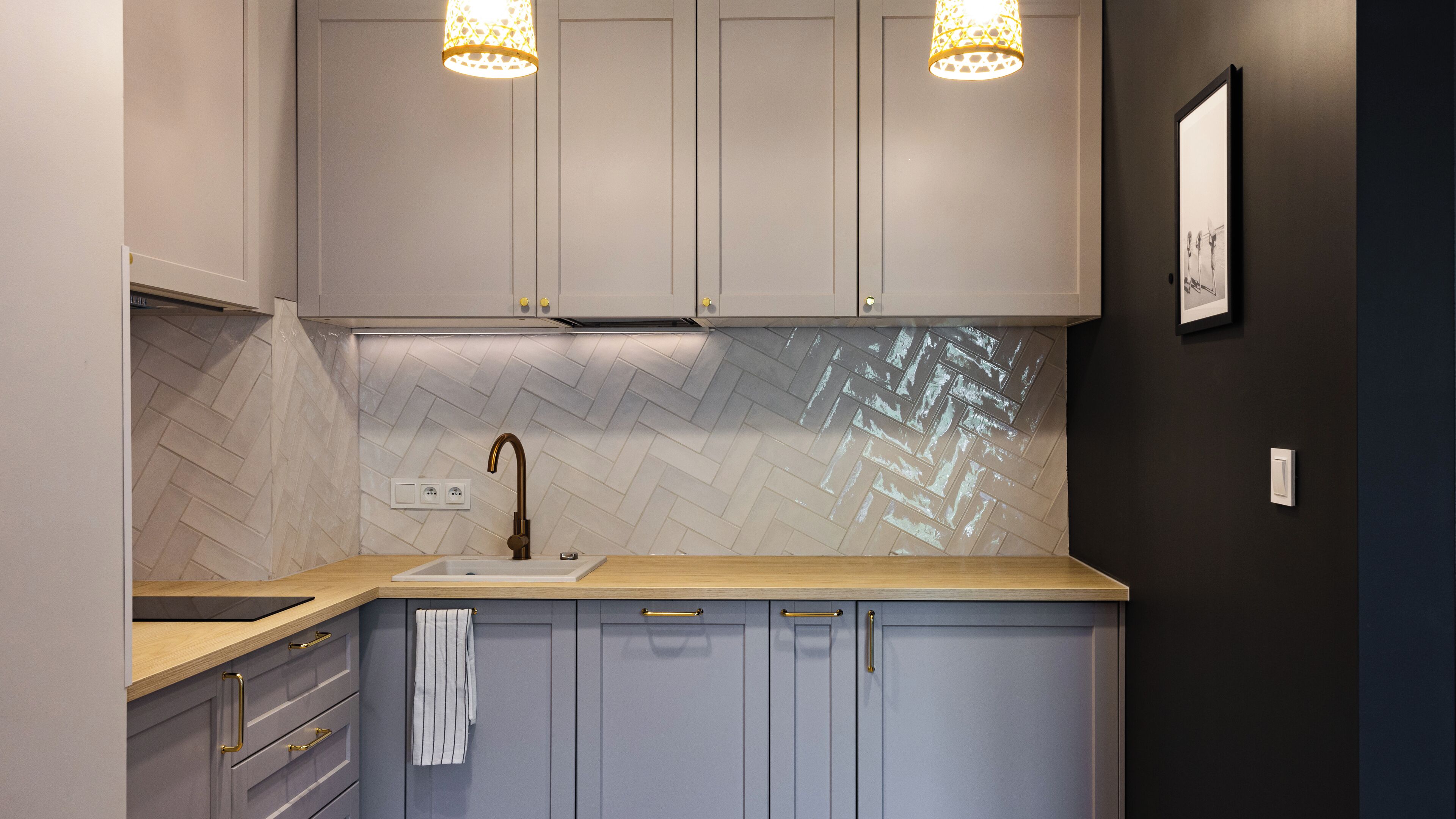 A modern kitchen with grey cabinets, a herringbone tile backsplash, a wooden countertop, and a single gold faucet.