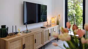 Ein modernes Wohnzimmer mit einem flachbildfernseher an der Wand, einem Holztisch mit Dekoration und einem großen Fenster mit Blick auf Grünflächen.