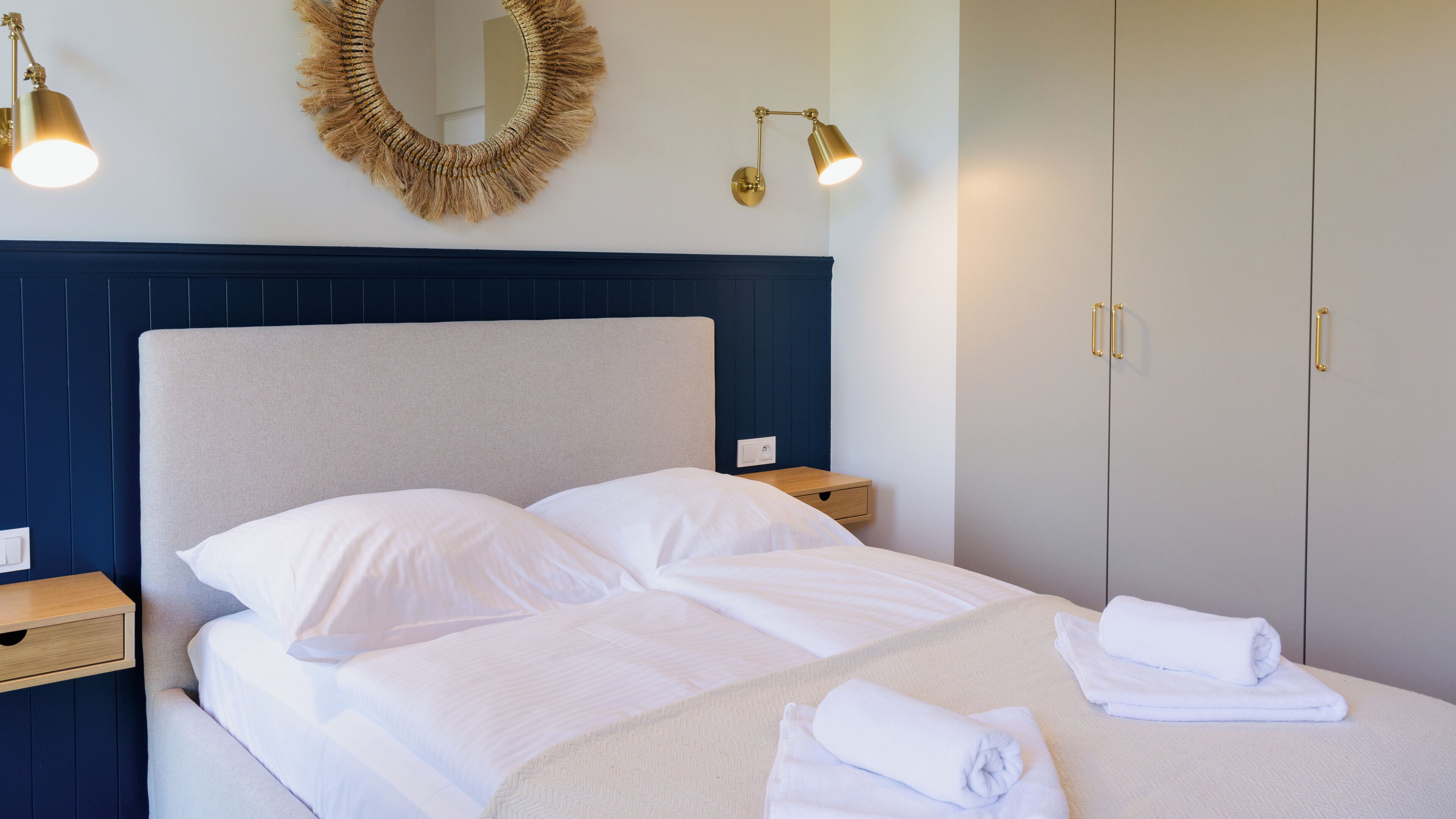 Una cama bien hecha con ropa de cama blanca, cabecera, lámparas de noche y un espejo en la pared.
