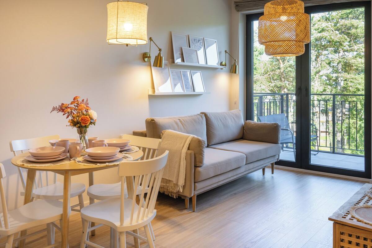 A modern living room with a dining area, featuring a sofa, chairs, a table set for dining, and a balcony with a view of greenery.