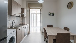 A modern kitchen with a built-in washing machine, a dining area with a wooden table and chairs, and a large window providing natural light. - Casa Adda by Wonderful Italy - Plutone (Syracuse)