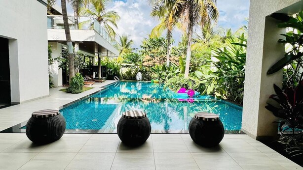 A swimming pool with three round black stools in the foreground, surrounded by lush greenery and palm trees. - Narayani Revtree - Resort & Villa (Hospet)