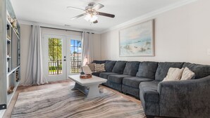 A living room with a gray sectional sofa, a white coffee table, a patterned rug, a ceiling fan, and a large window with a view of greenery. - Walk to Beach: 7BR House w/ Private Pool & Hot Tub (Surfside Beach)