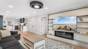 A modern living room with a flat-screen TV, a fireplace, and a kitchen in the background. - Walk to Beach: 7BR House w/ Private Pool & Hot Tub (Surfside Beach)