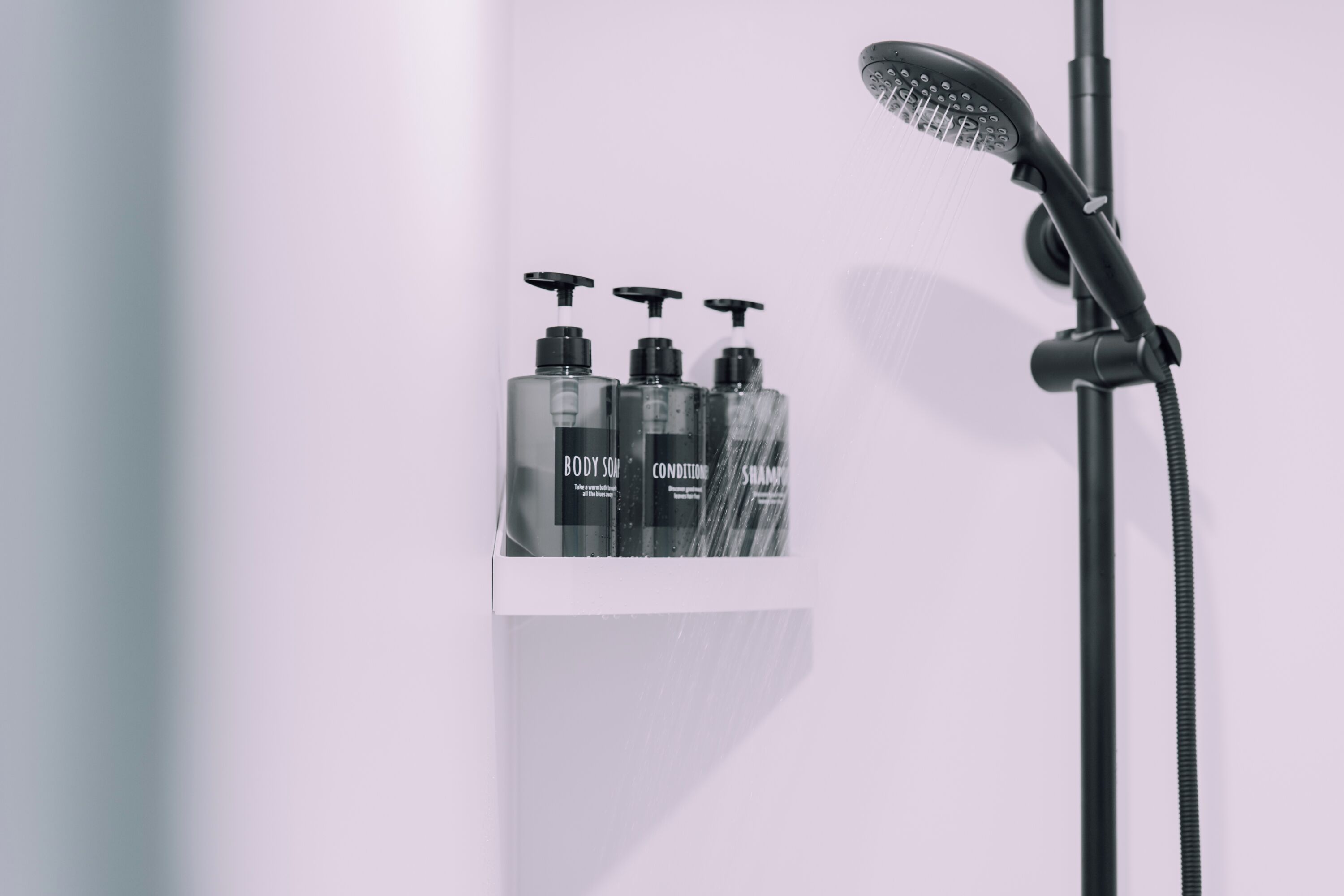Un espace douche équipé d’un pommeau de douche à main et de trois flacons de savon pour le corps étiquetés « CORPS », « SOIN » et « SHAMPOING ».