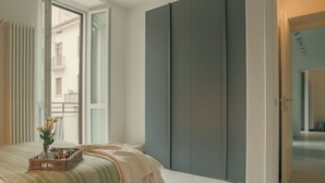 Et soveværelse med en seng, et natbord med en vase og stearinlys, en grå garderobe, og udsigt til altanen.