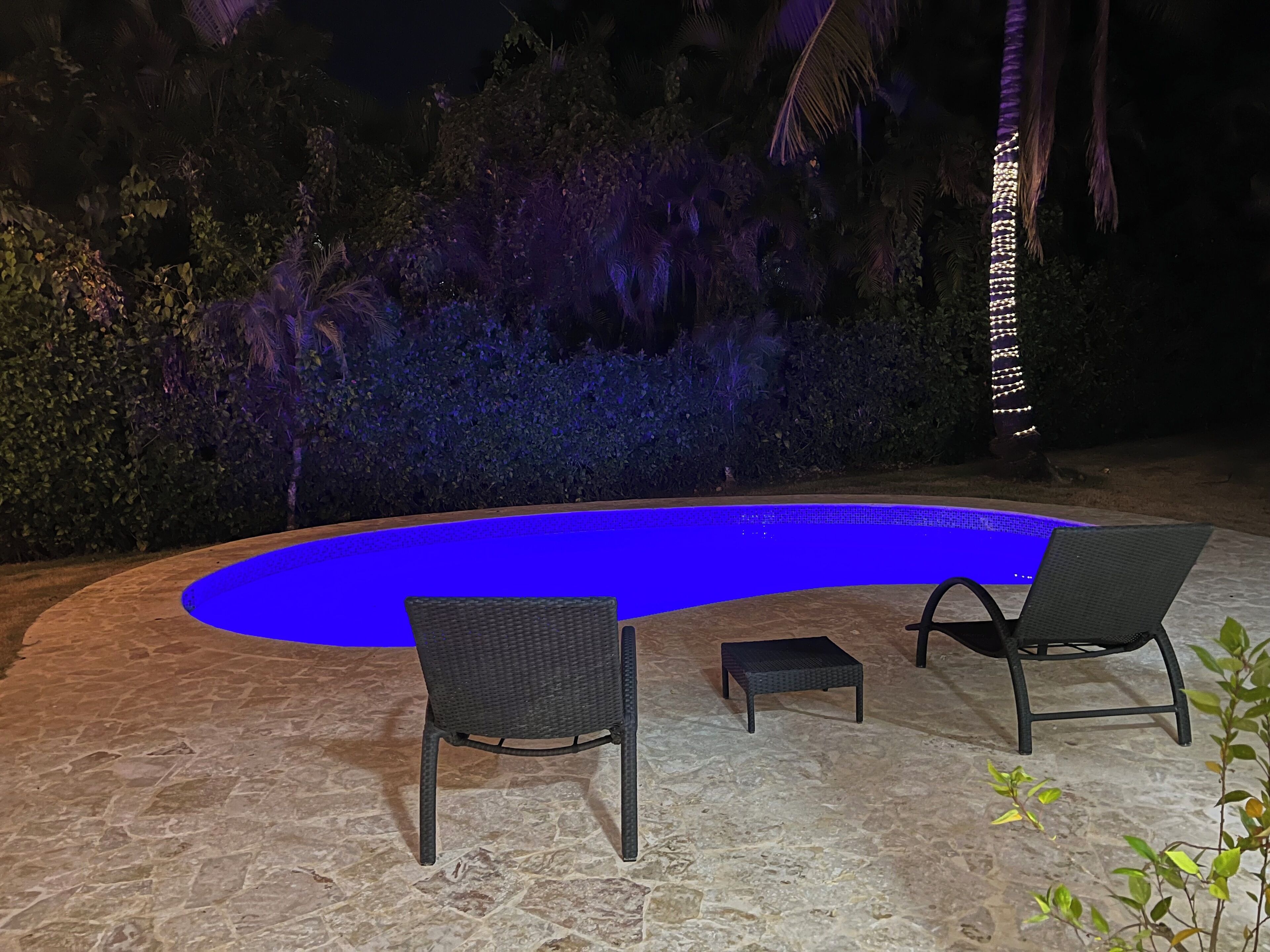 A swimming pool with a blue-lit edge, surrounded by stone flooring, two wicker chairs, and a small table.