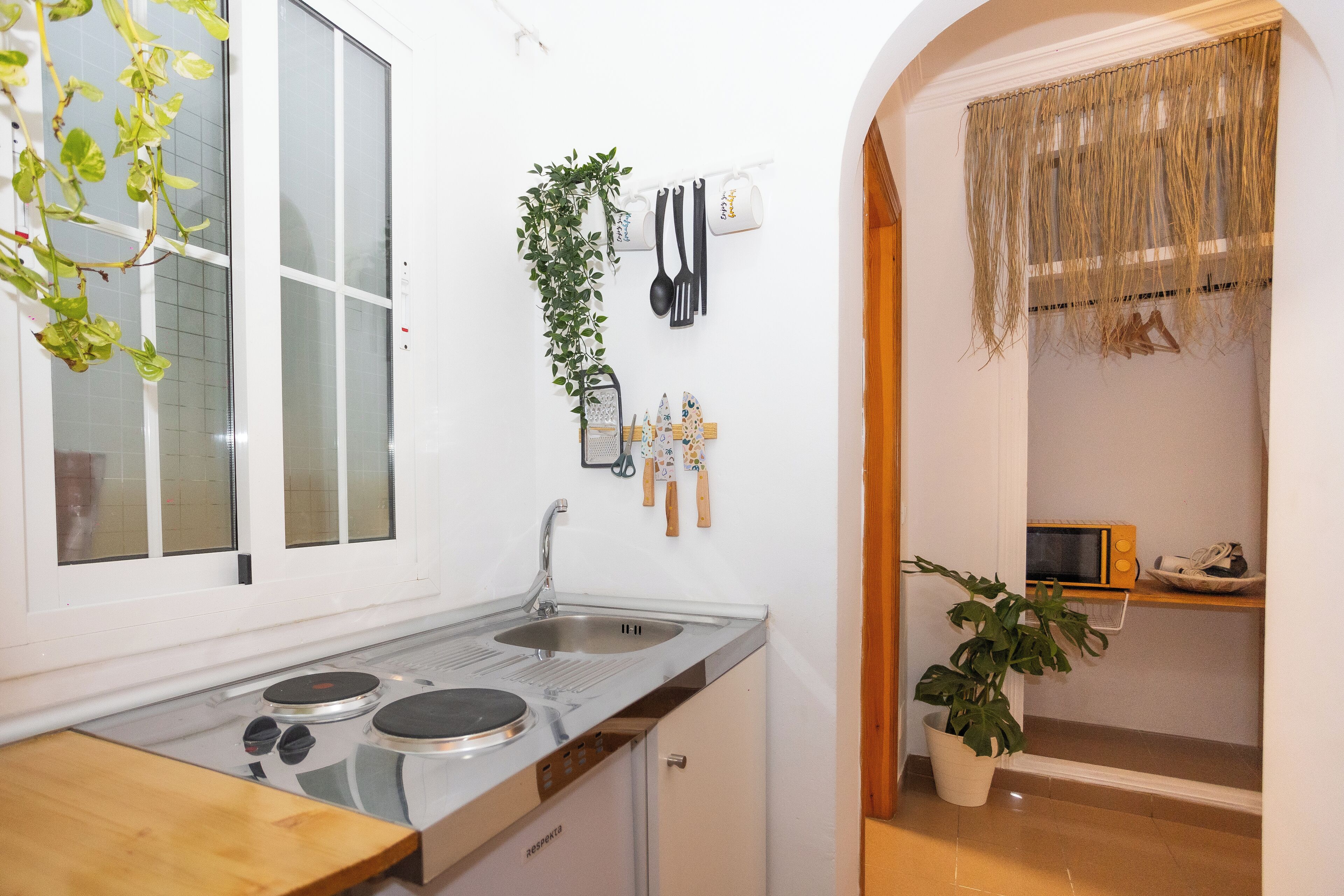 A compact kitchen with a sink, stove, and a small countertop. There are plants and kitchen utensils on the wall.