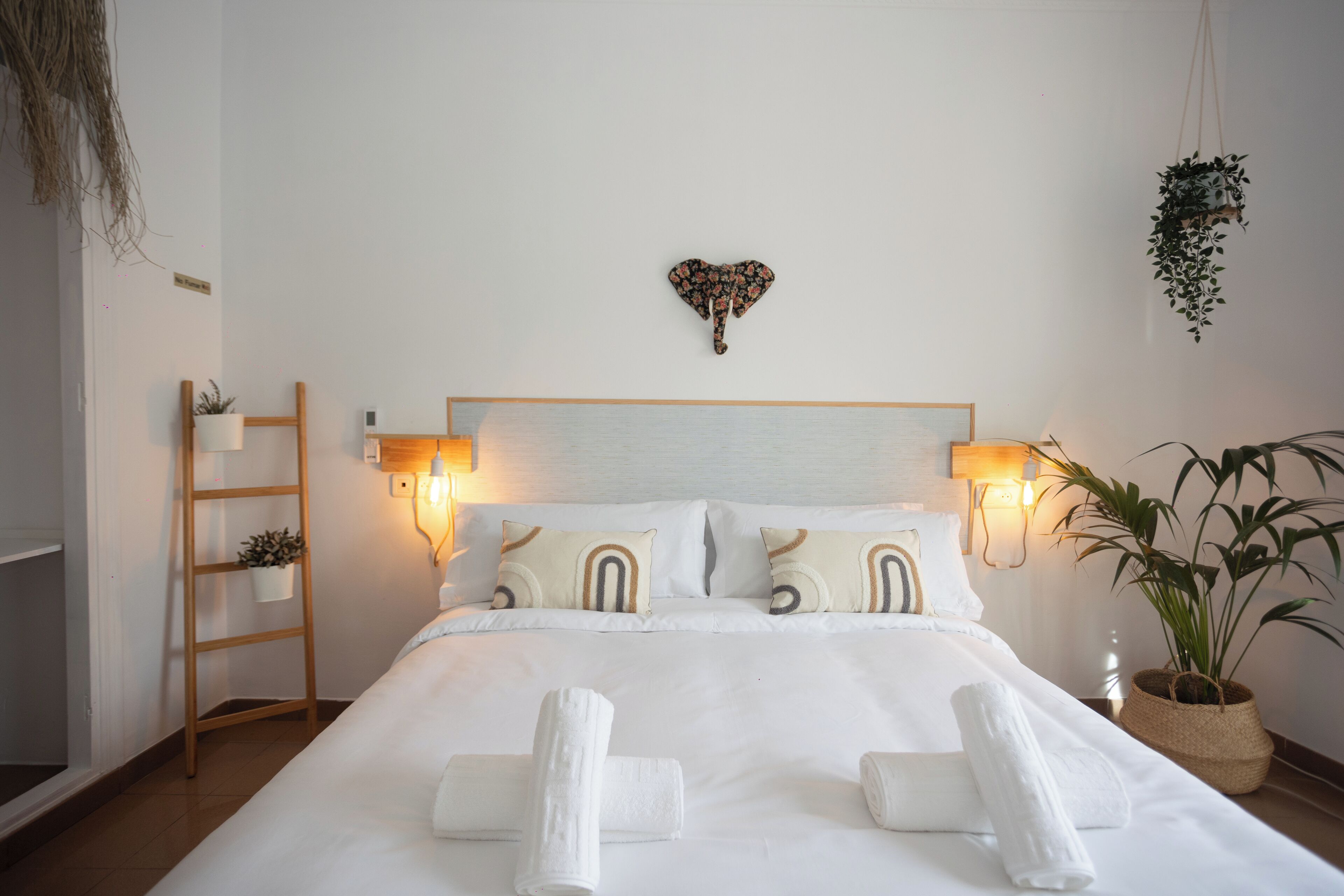 Una cama bien tendida con sábanas y toallas blancas, una escalera de madera y una planta en la pared.