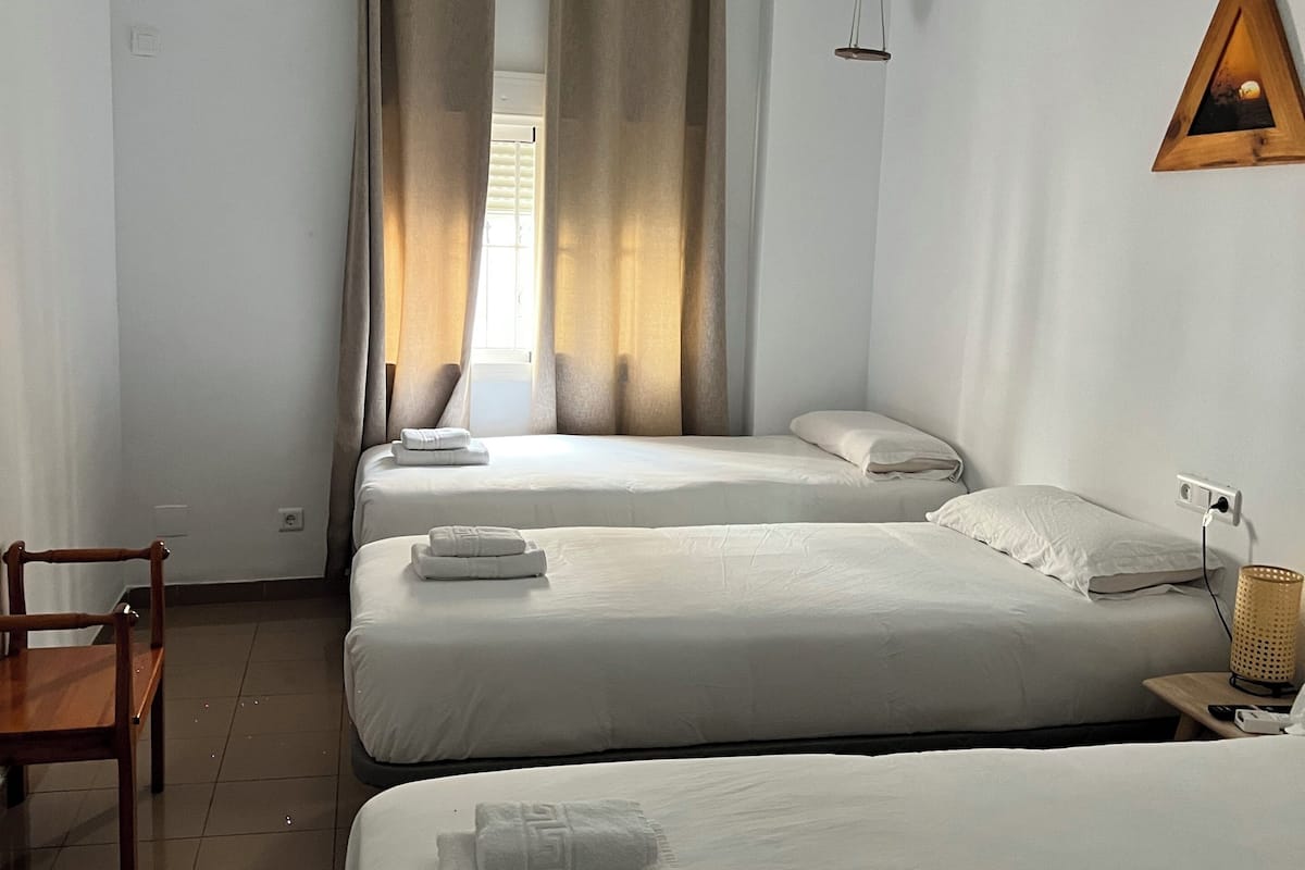 A hotel room with two beds, a wooden chair, a small table, and a wall-mounted lamp.