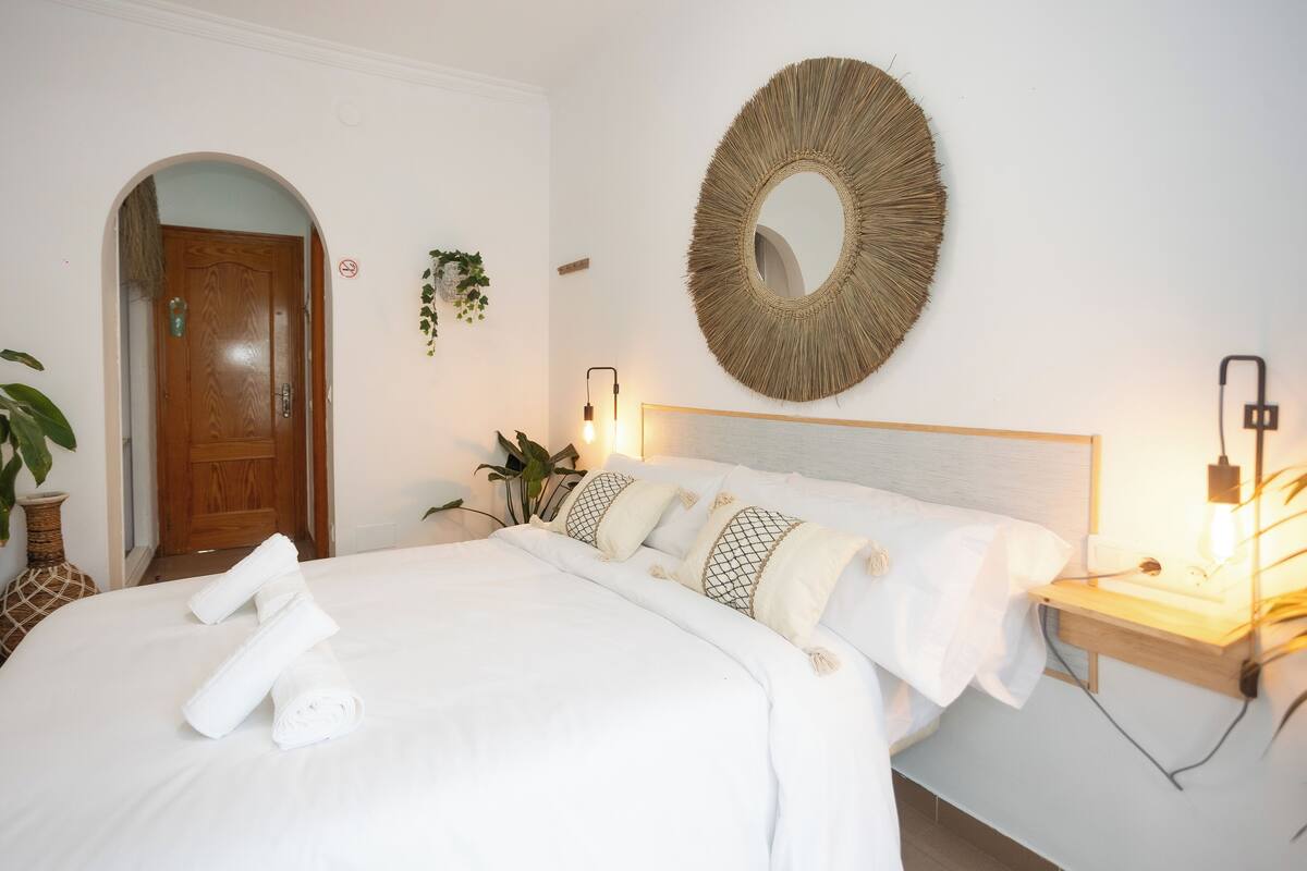 A neatly made bed with white linens and rolled towels, a round woven mirror on the wall, and a bedside table with a lamp.