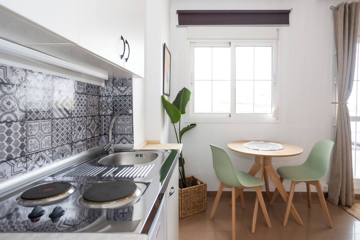 A kitchen with a modern stove, a sink, and patterned backsplash tiles. There is a dining table with two chairs and a potted plant.