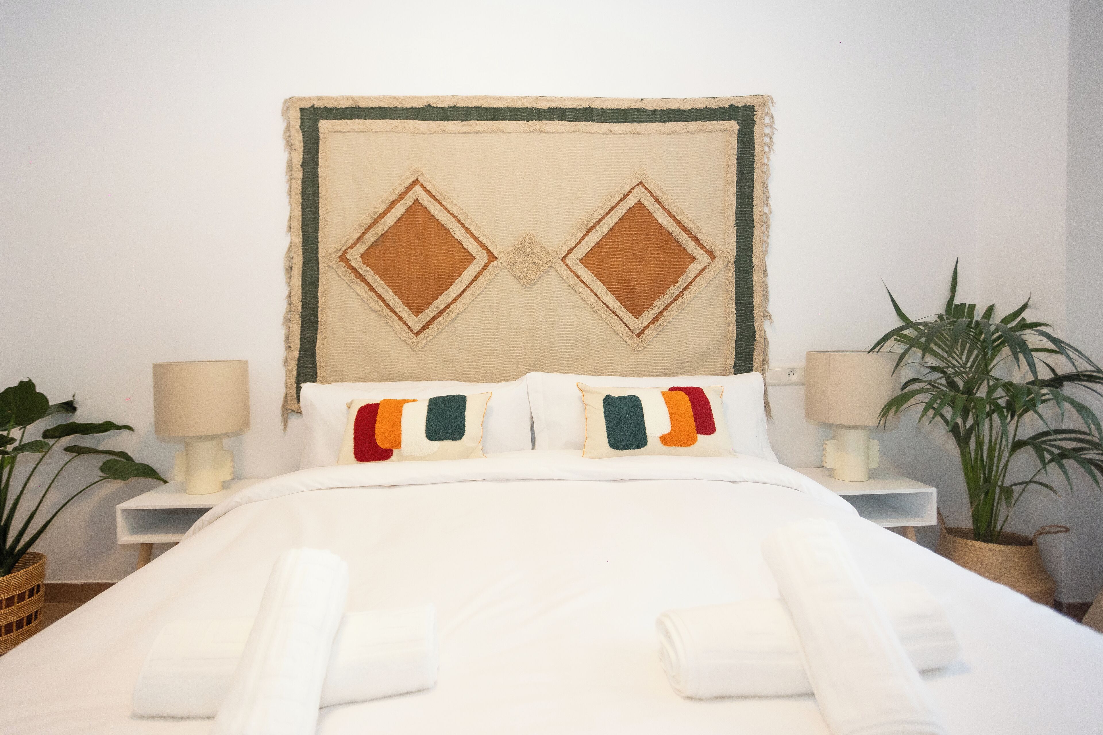Una cama bien tendida con sábanas blancas y almohadas de colores, un cabecero con diseño geométrico y dos mesitas de noche con lámparas y plantas en macetas.