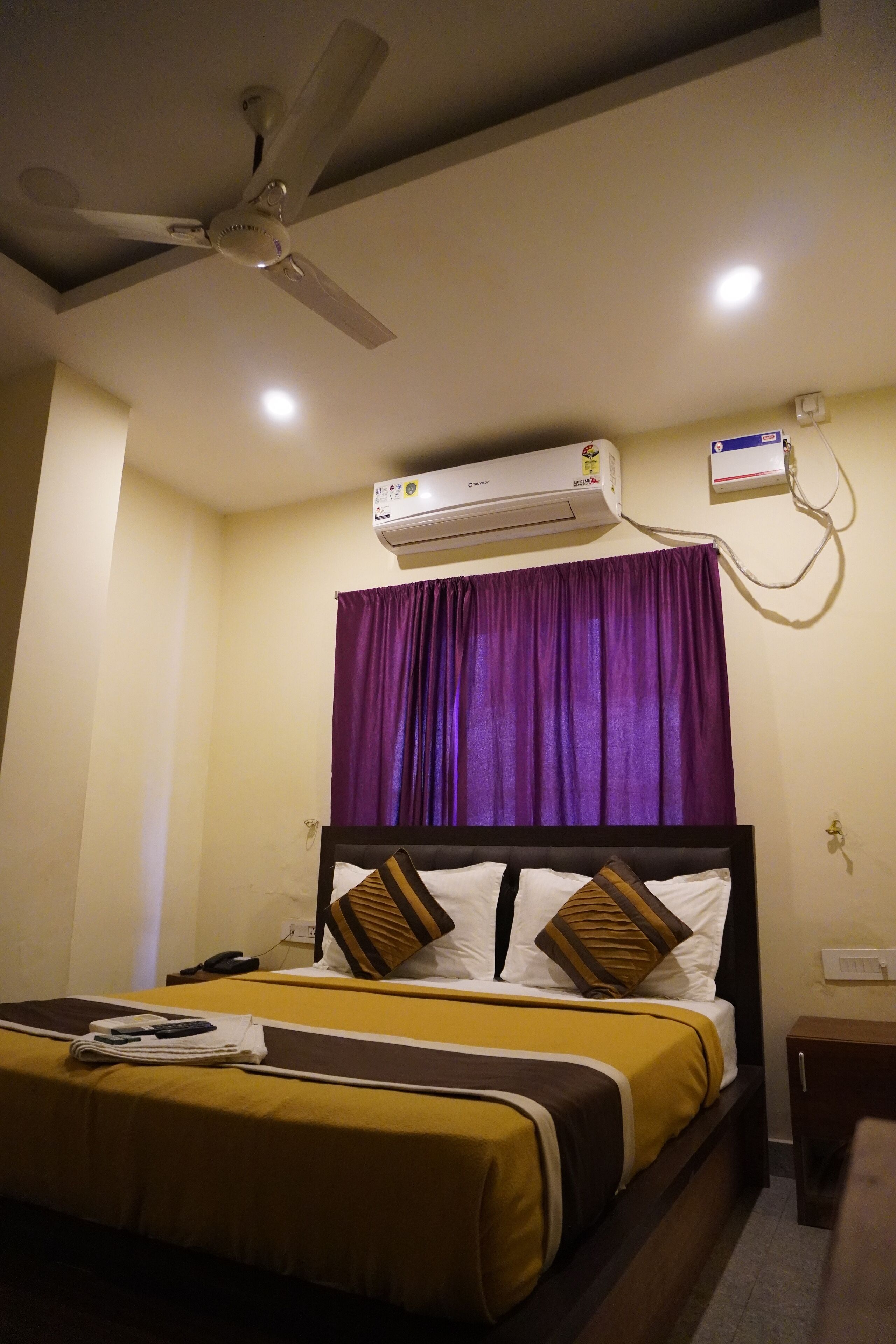 A hotel room with a bed, a ceiling fan, an air conditioning unit, and a nightstand.