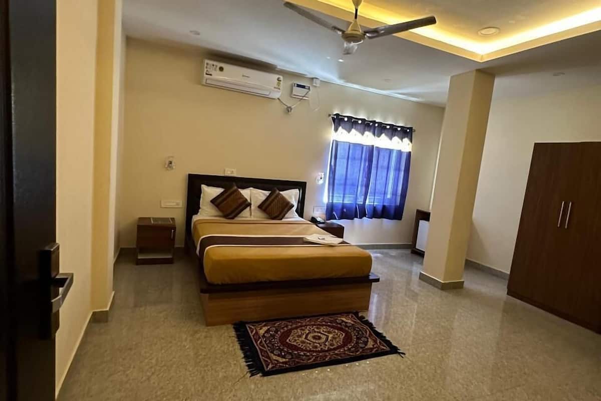 A hotel room with a bed, a ceiling fan, an air conditioning unit, a window with curtains, and a wooden wardrobe.