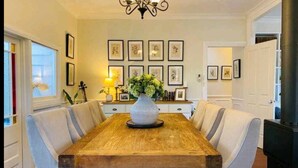 A dining room with a wooden table, light blue upholstered chairs, a vase with flowers, framed artwork on the wall, and a fireplace. - The Hollies (Monbulk)