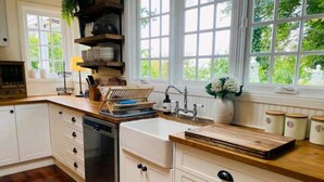 A kitchen with white cabinets, a dishwasher, a wooden countertop, and a window with a view of greenery. - The Hollies (Monbulk)