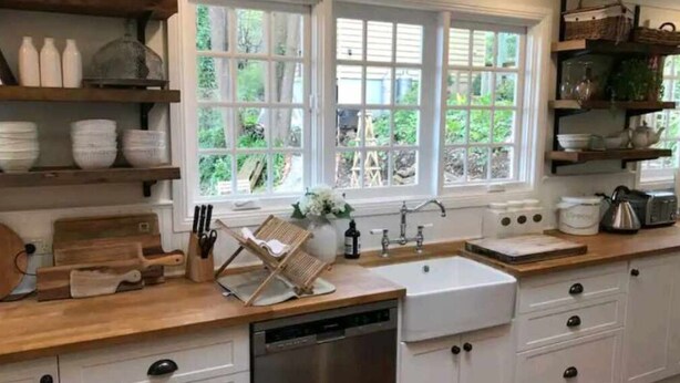A kitchen with white cabinets, a wooden countertop, a dishwasher, a sink, and a window with a view of greenery. - The Hollies (Monbulk)