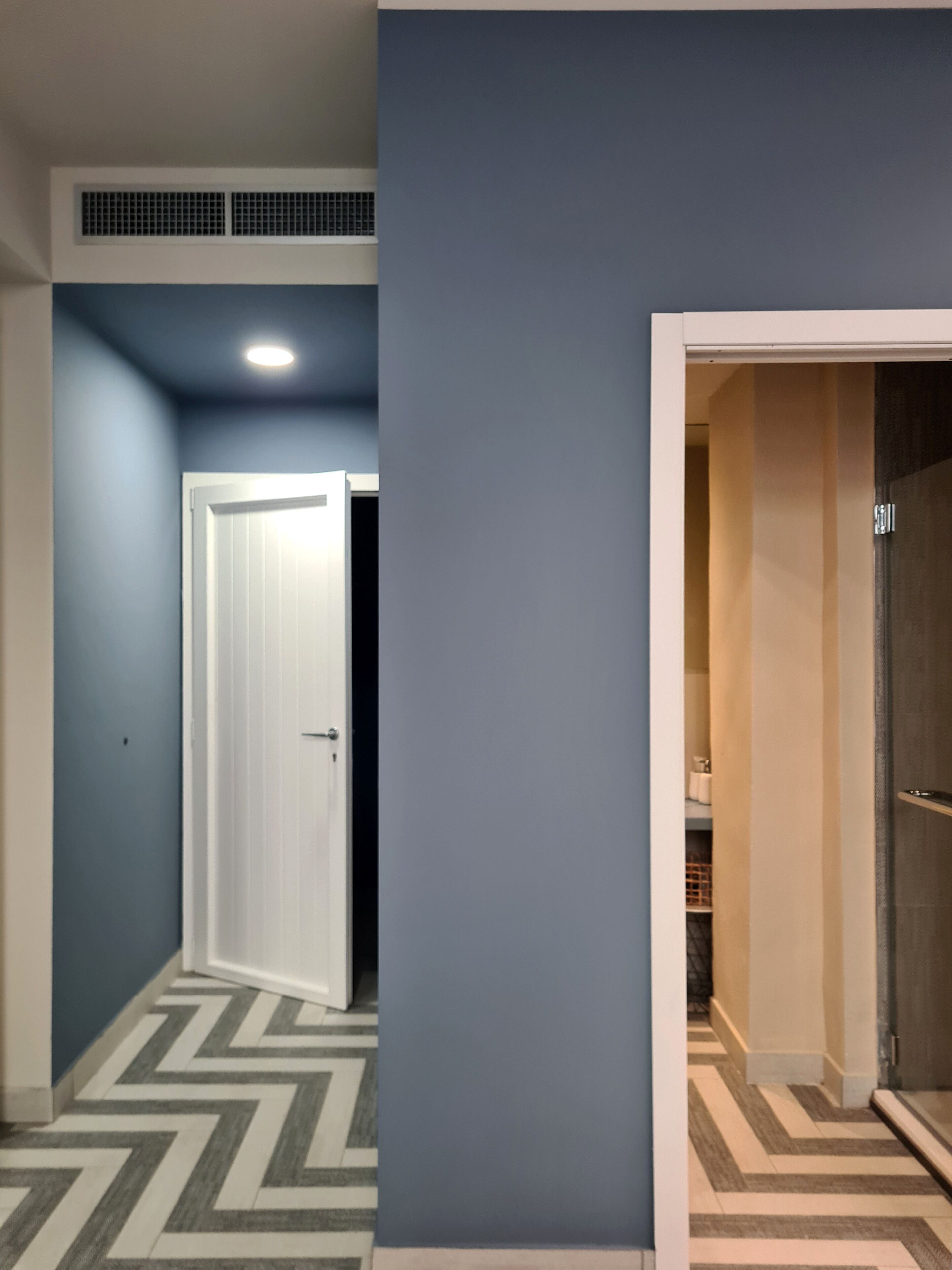 Un couloir avec une porte blanche, une grille d’aération au plafond et un revêtement de sol à motifs.