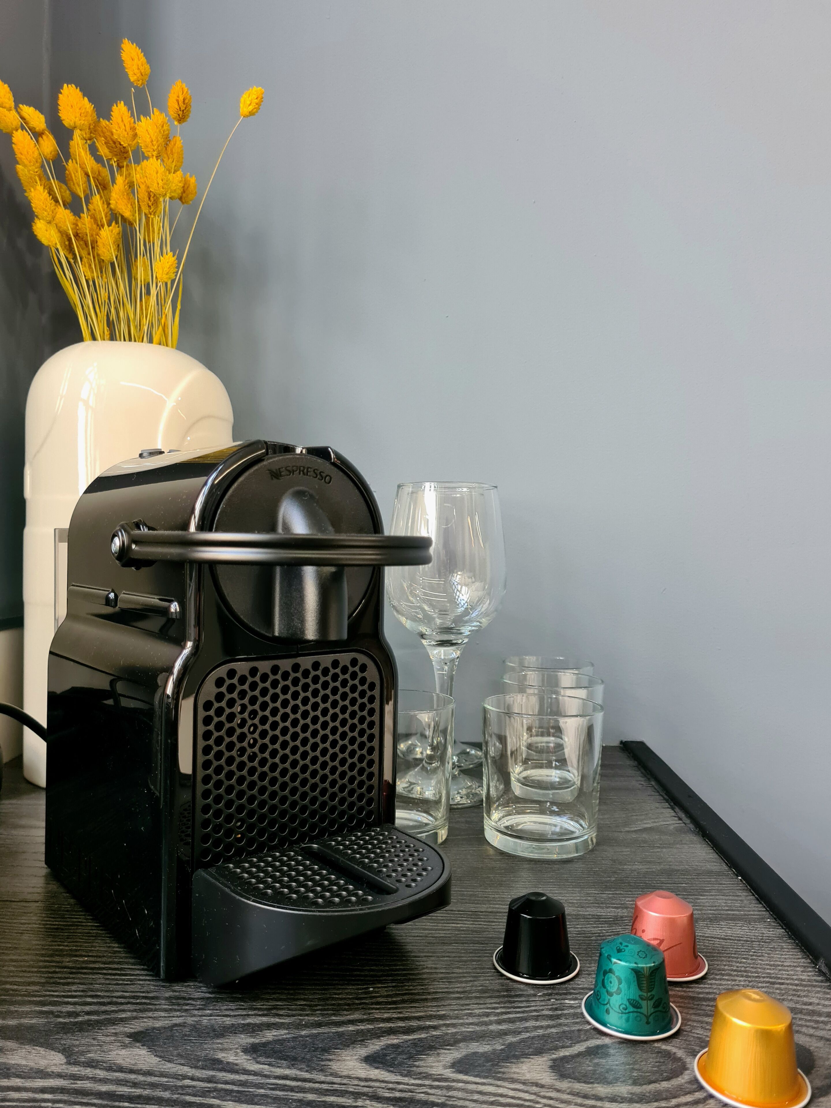 A coffee machine with a glass container, a vase with yellow flowers, and several empty glasses on a wooden surface.
