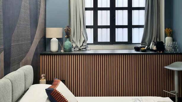 A modern hotel room with a large bed, a window with curtains, a bedside table with a lamp and decorative items, and a view of the city through the window. - Serendipity Rooms - Navigli (Milan)