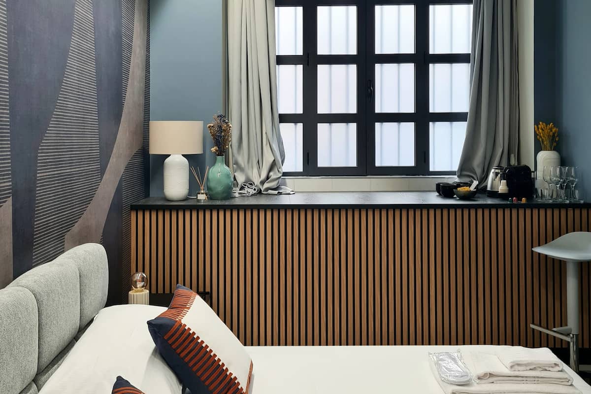 A modern hotel room with a large bed, a window with curtains, a bedside table with a lamp and decorative items, and a view of the city through the window.