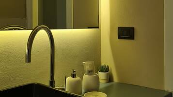Un bagno moderno con un lavandino nero, uno specchio, una lampada a parete e una piccola pianta in vaso.