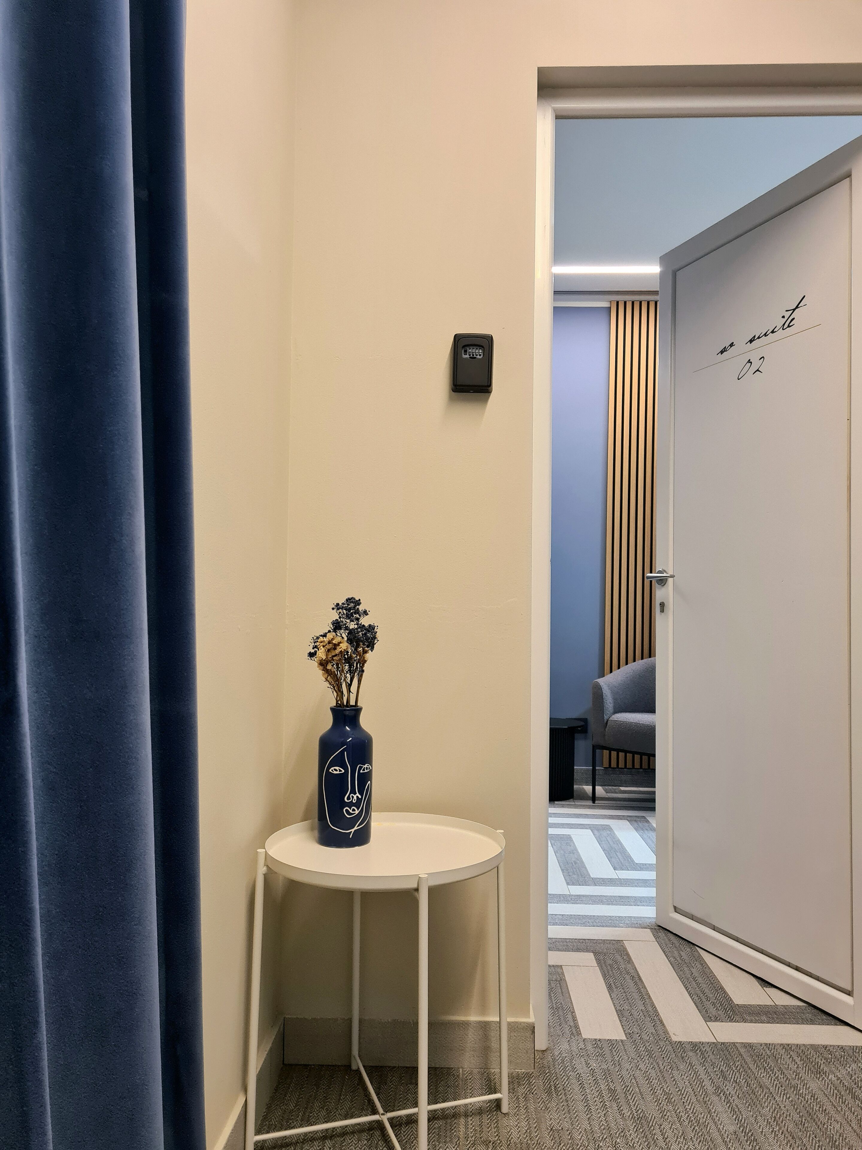 A hotel room with a door marked "no suite 02", a small table with a vase of flowers, and a blue curtain.