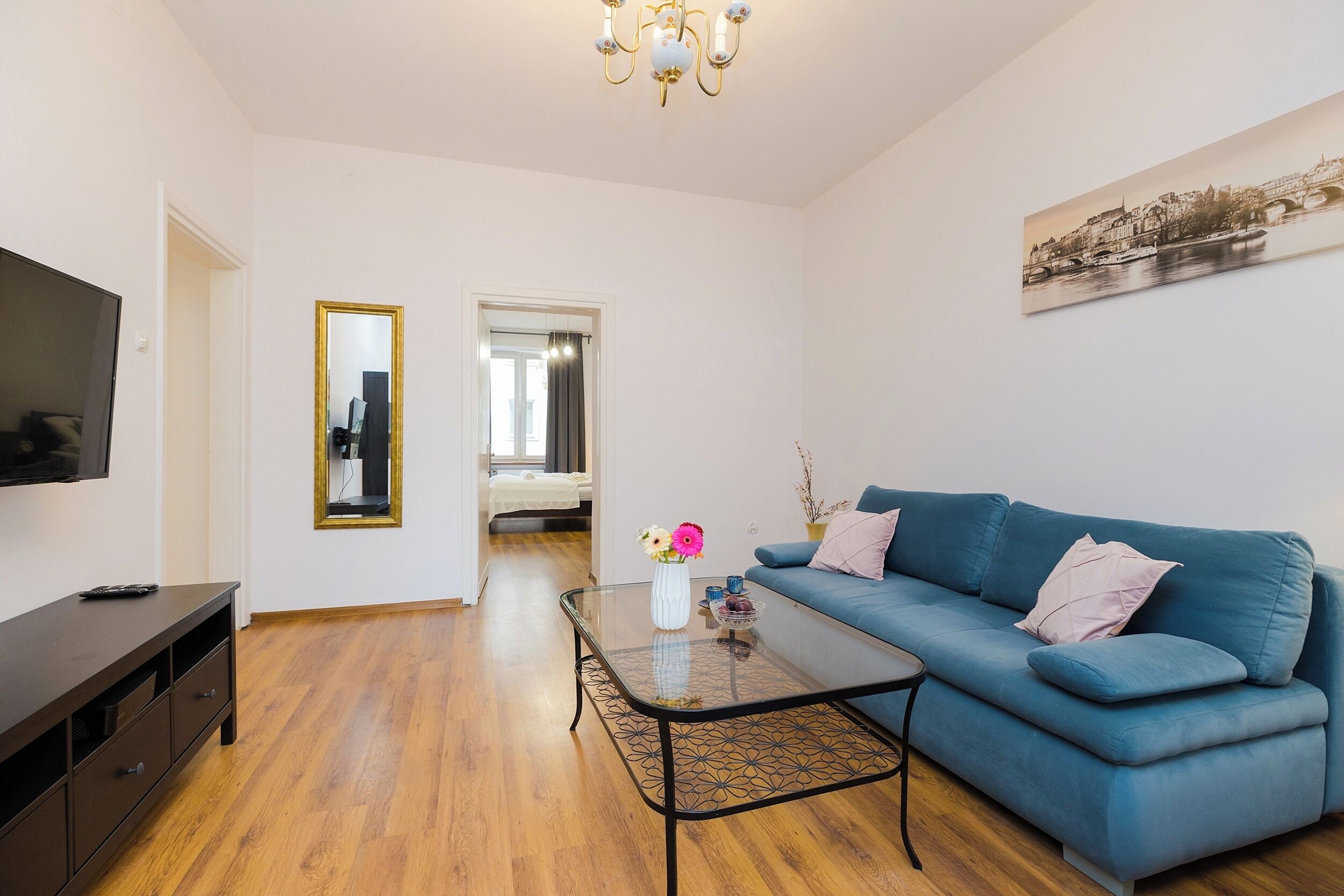A living room with a blue sofa, a glass coffee table, a TV on a stand, and a painting on the wall.