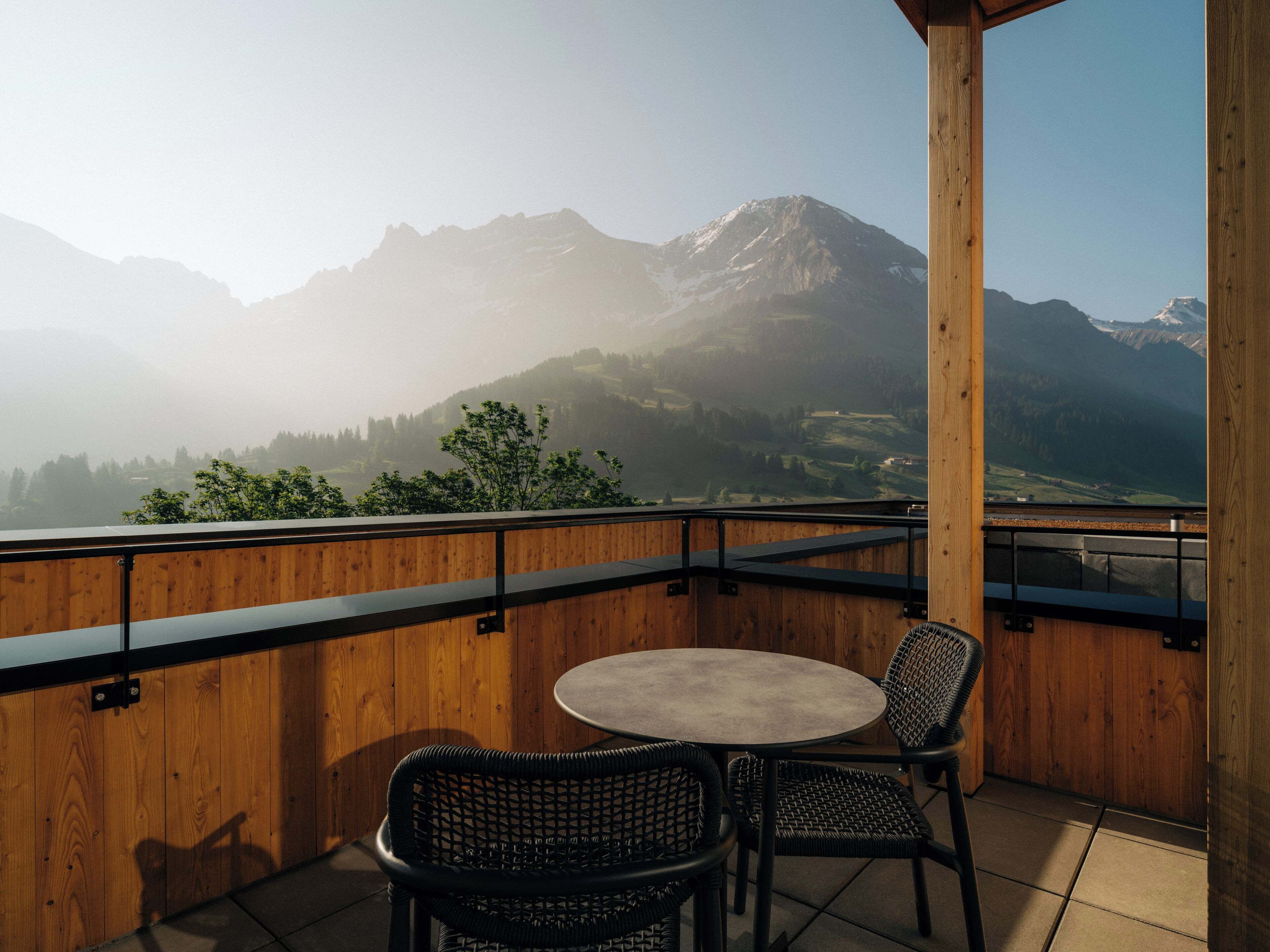 A wooden balcony with a round table and two wicker chairs, overlooking a mountainous landscape.