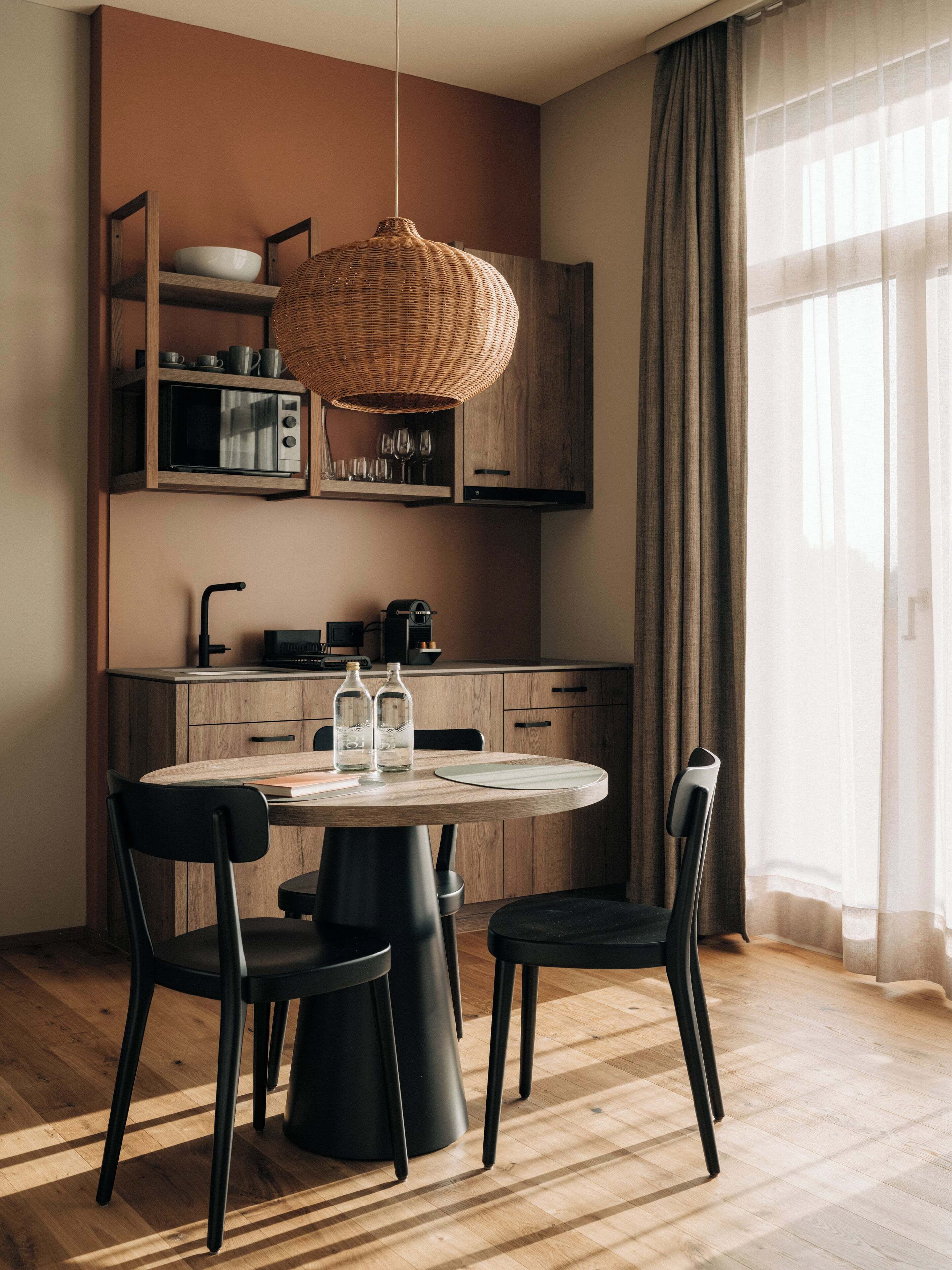 A modern kitchen with a round dining table, black chairs, a hanging light fixture, and wooden cabinetry.