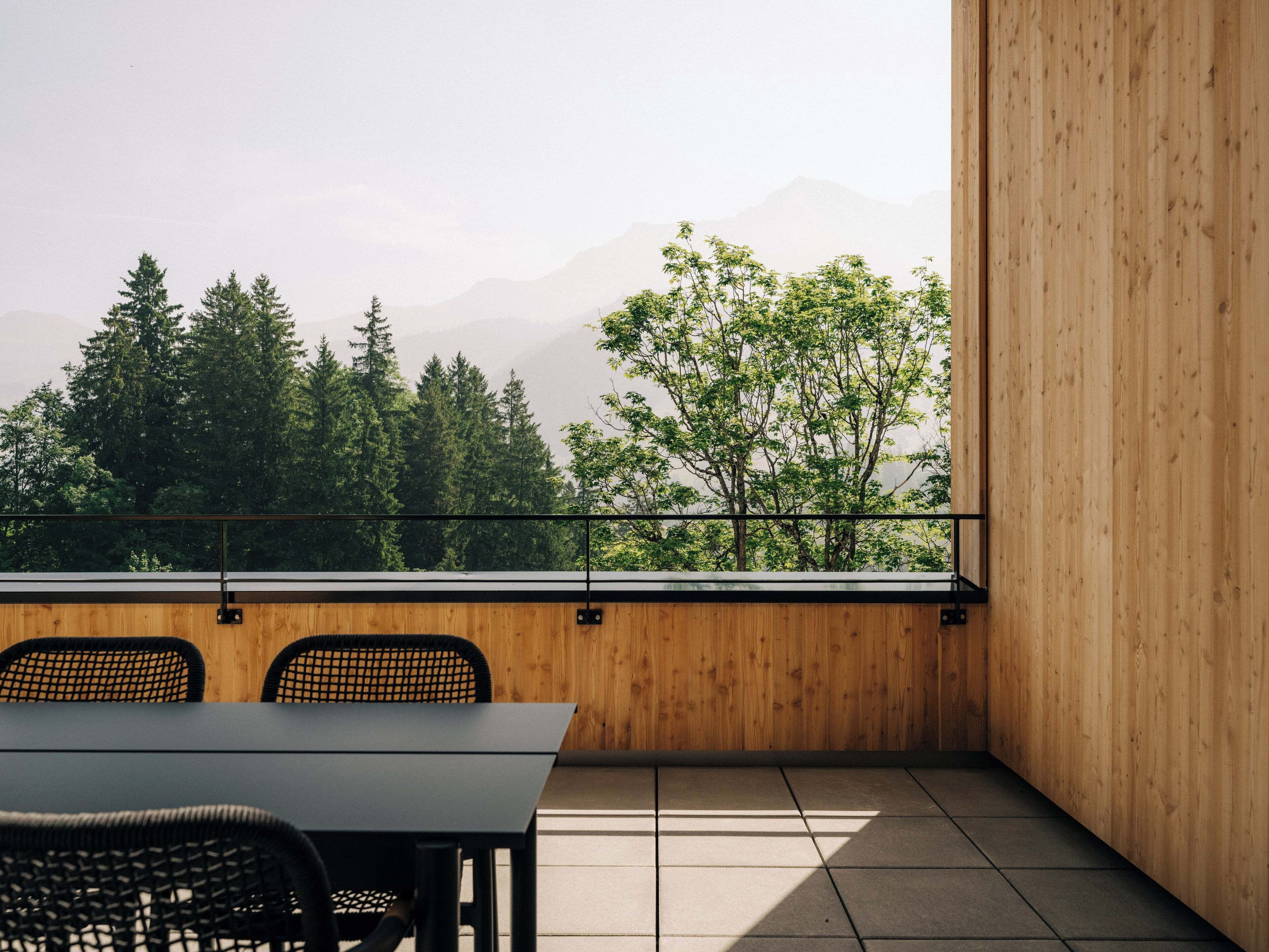 Quarto moderno com piso de madeira, uma mesa e duas cadeiras. Possui uma janela grande com vista para árvores e montanhas.