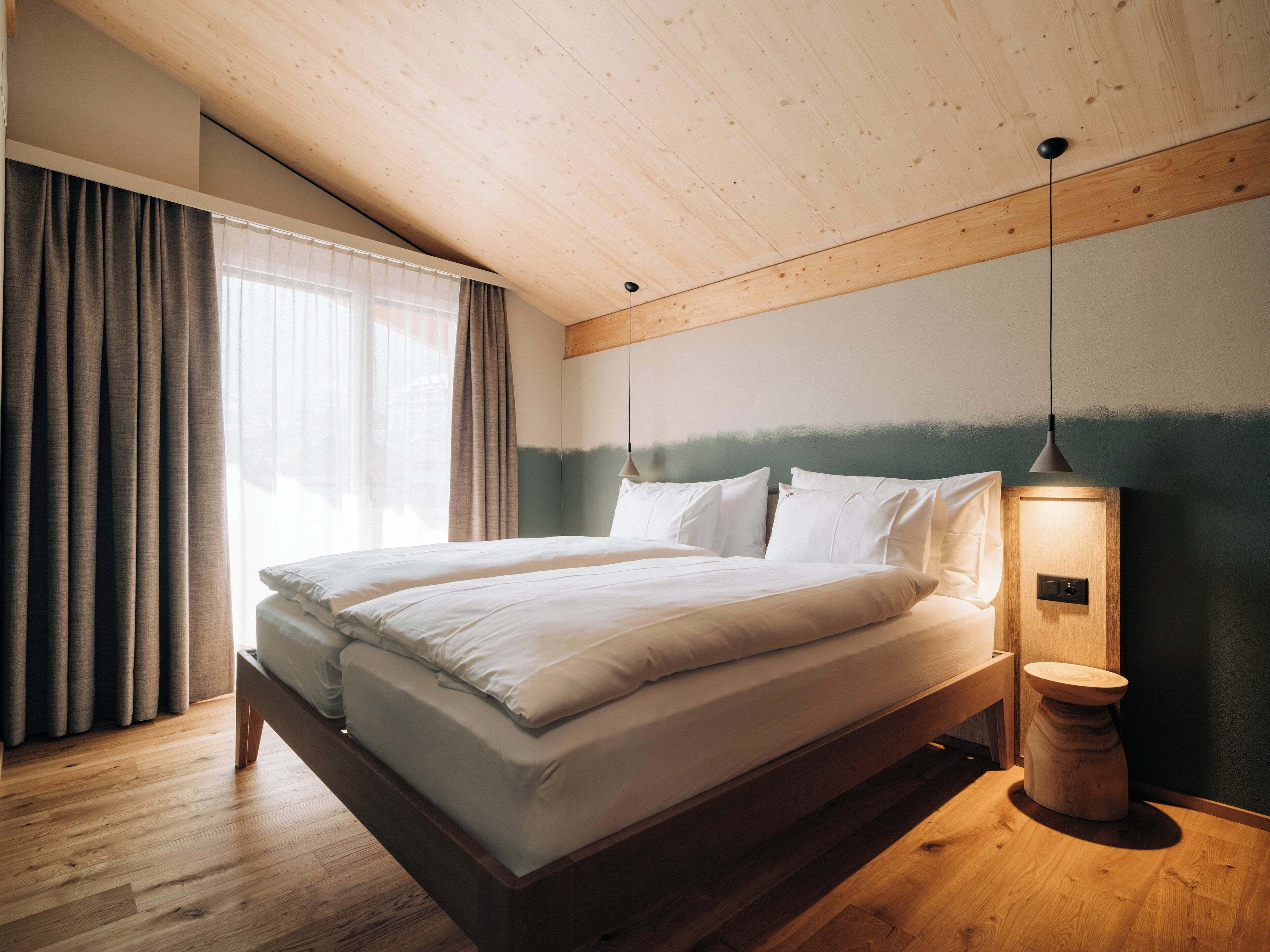 Une chambre moderne avec un plafond en bois, un grand lit avec une literie blanche, une table de chevet avec une lampe, et une vue sur l’extérieur par une fenêtre agrémentée de rideaux.