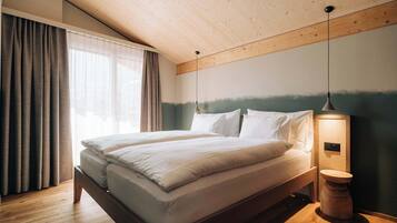 Une chambre moderne avec un plafond en bois, un grand lit avec une literie blanche, une table de chevet avec une lampe, et une vue sur l’extérieur par une fenêtre agrémentée de rideaux.