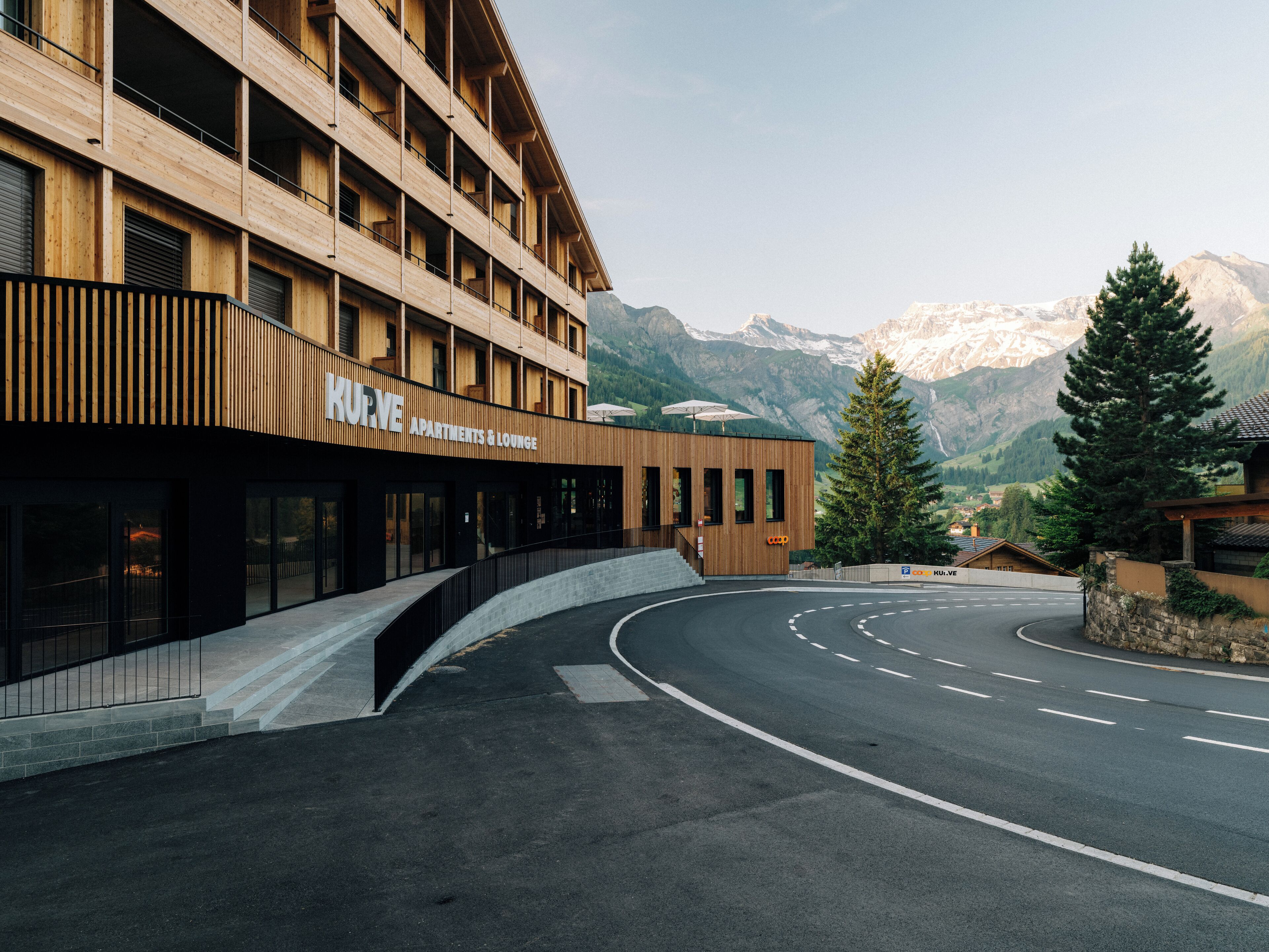 A modern building with a wooden facade, labeled "KURVE Apartments & Lounge," featuring a curved driveway and a mountain backdrop.