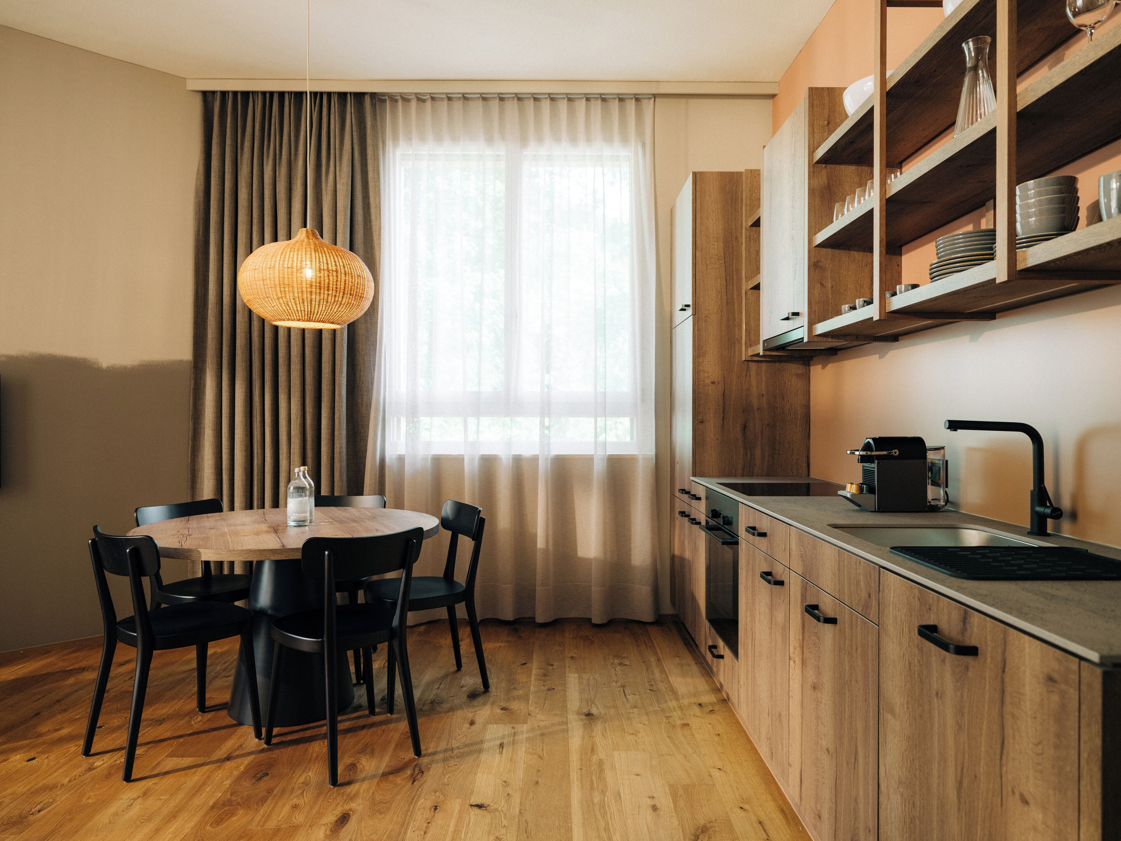 Une cuisine de taille réduite avec des meubles en bois, une table à manger avec des chaises, un luminaire suspendu et une fenêtre avec des rideaux.