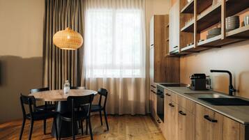 Une cuisine de taille réduite avec des meubles en bois, une table à manger avec des chaises, un luminaire suspendu et une fenêtre avec des rideaux.