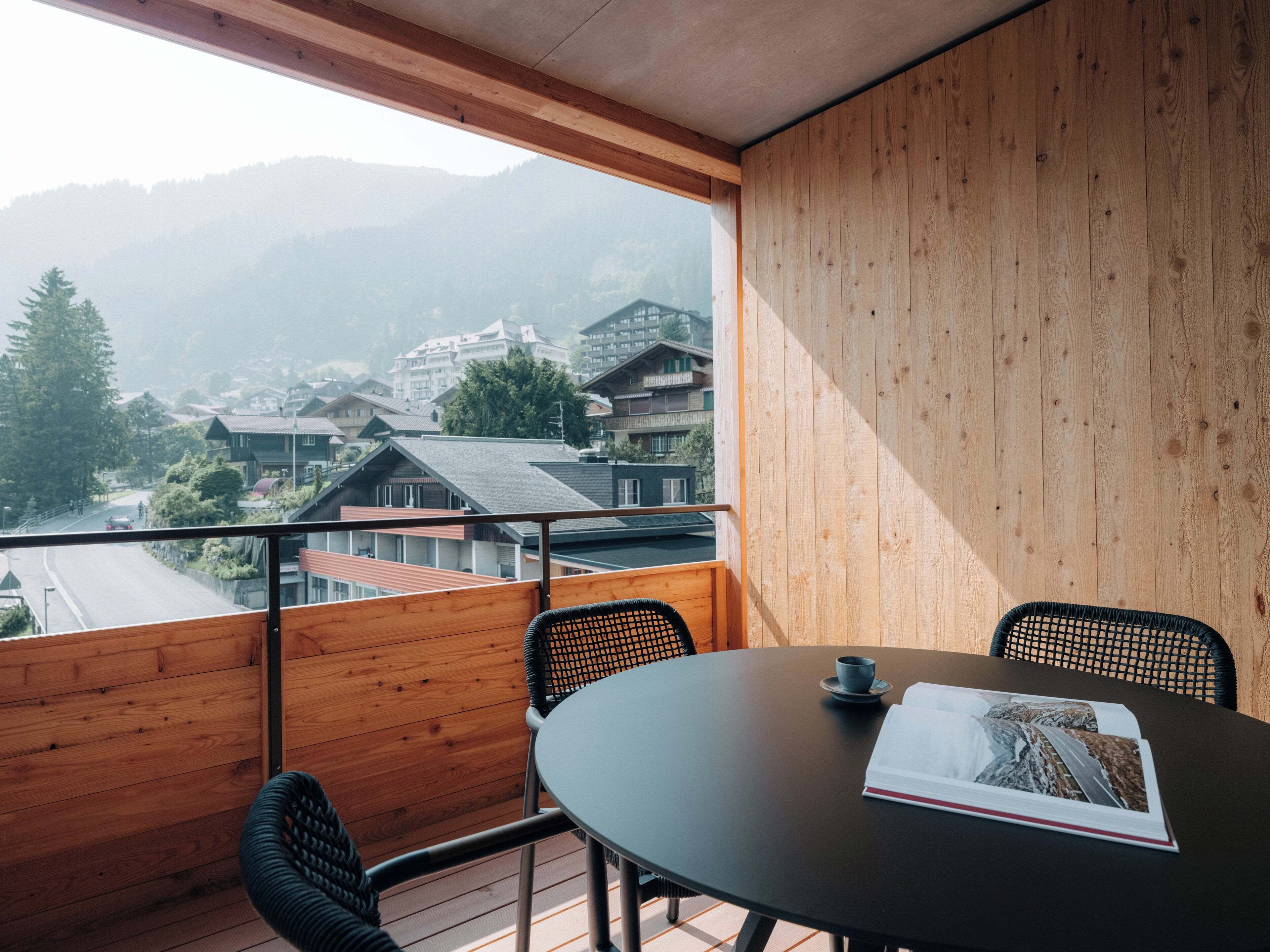 Un balcón con piso de madera, una mesa redonda, dos sillas y vista a un pueblo con edificios y montañas al fondo.