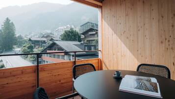 Un balcón con piso de madera, una mesa redonda, dos sillas y vista a un pueblo con edificios y montañas al fondo.