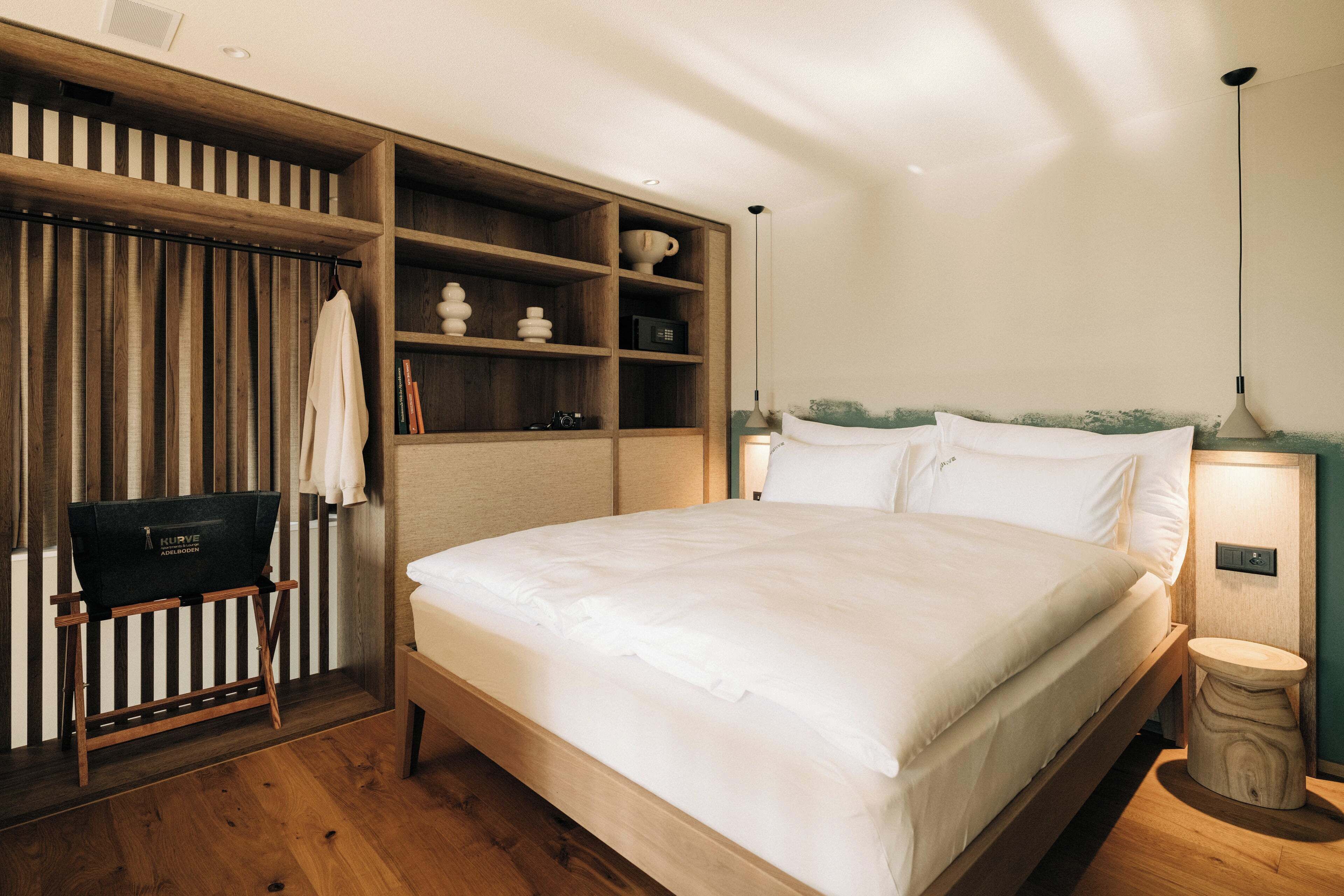 A bedroom with a wooden bed frame, a built-in wardrobe, a small seating area with a chair, and a bedside table with a lamp.